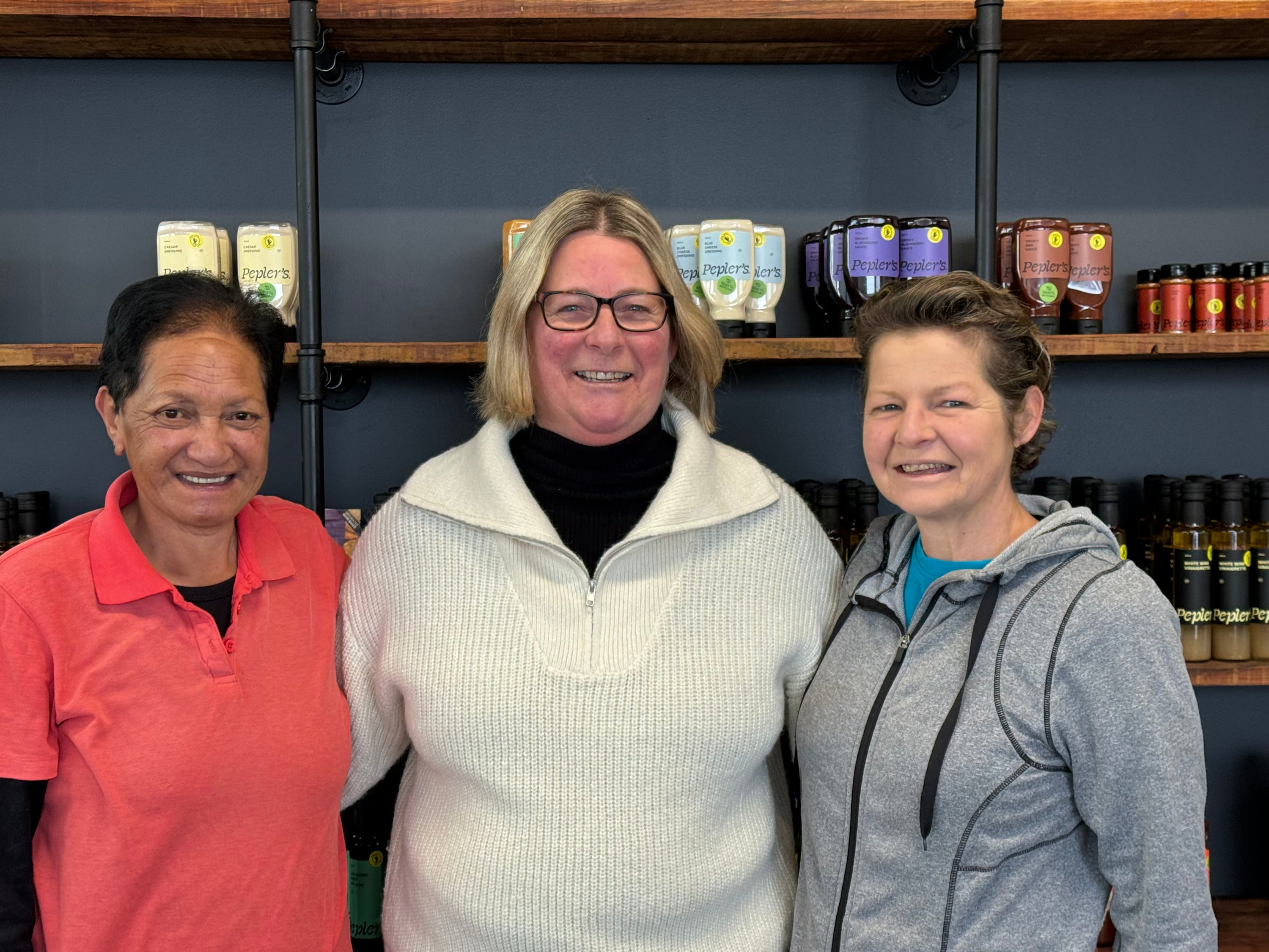 Three women standing together in front of a wooden shelf filled with various food products, including jars and bottles. The woman on the left wears a bright pink shirt, the woman in the middle is dressed in a white sweater and glasses, and the woman on the right is in a gray hoodie. They are smiling and appear to be in a welcoming, retail environment.