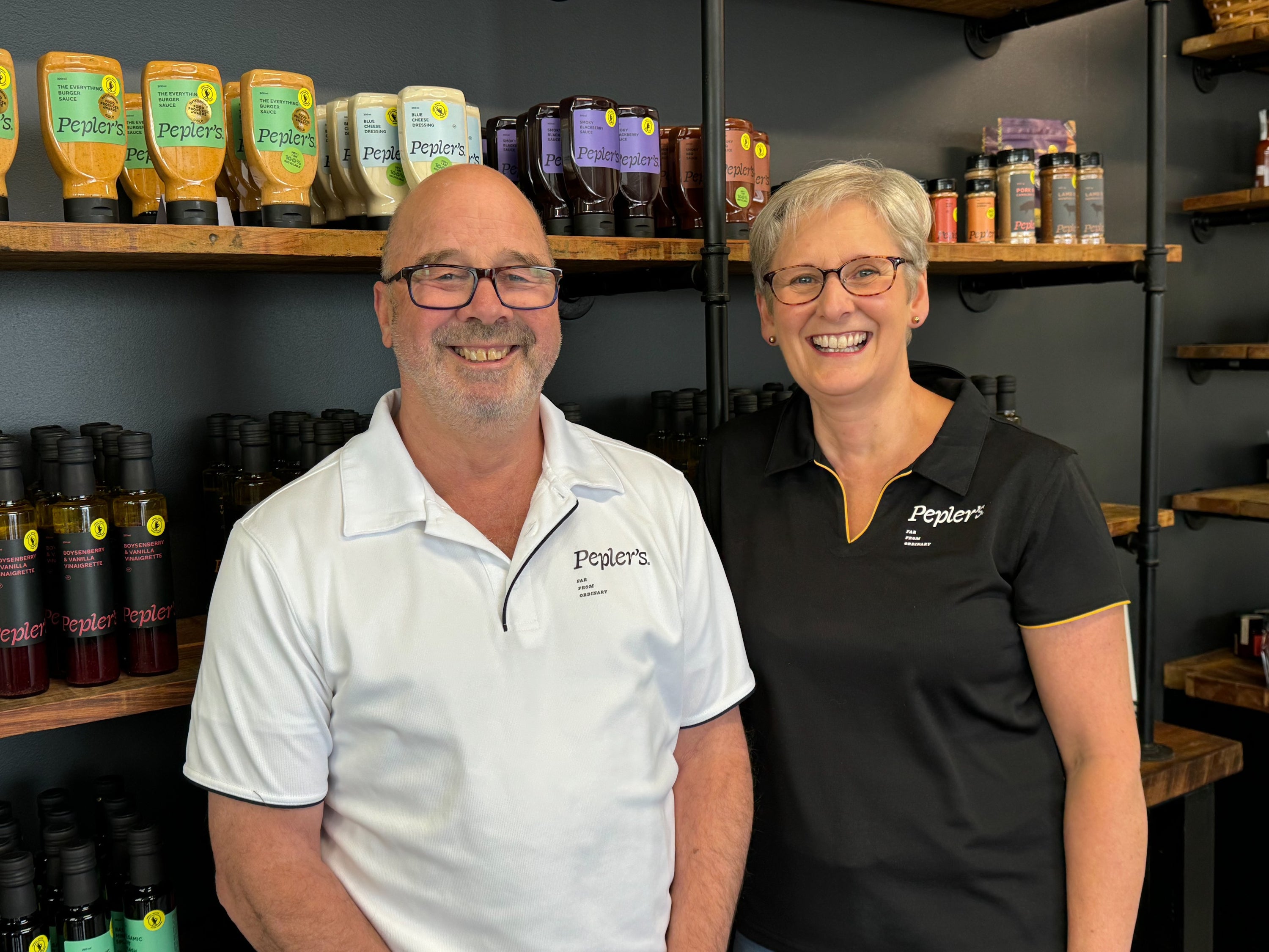 A smiling man, Paul, in a white shirt and a woman, Jane, in a black shirt with yellow accents stand in front of shelves stocked with various bottles and sauces labeled "Pepler's." The background features a dark wall and wooden shelves displaying colourful product packaging, creating a welcoming atmosphere.