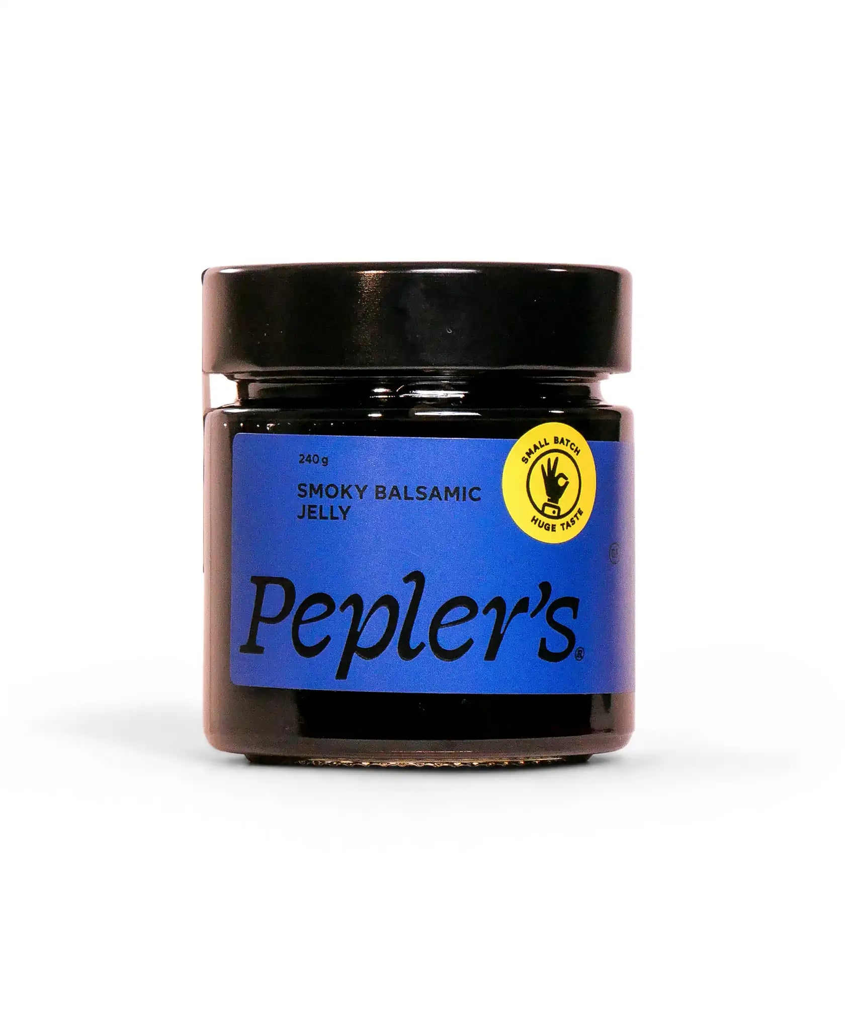 A glass jar of Pepler's Smoky Balsamic Jelly, weighing 240 grams, featuring a navy blue label with bold black lettering. The jar has a black lid and includes a yellow circular logo with a hand symbol, stating "Small Batch Huge Taste." The background is plain and light-coloured, emphasising the product.