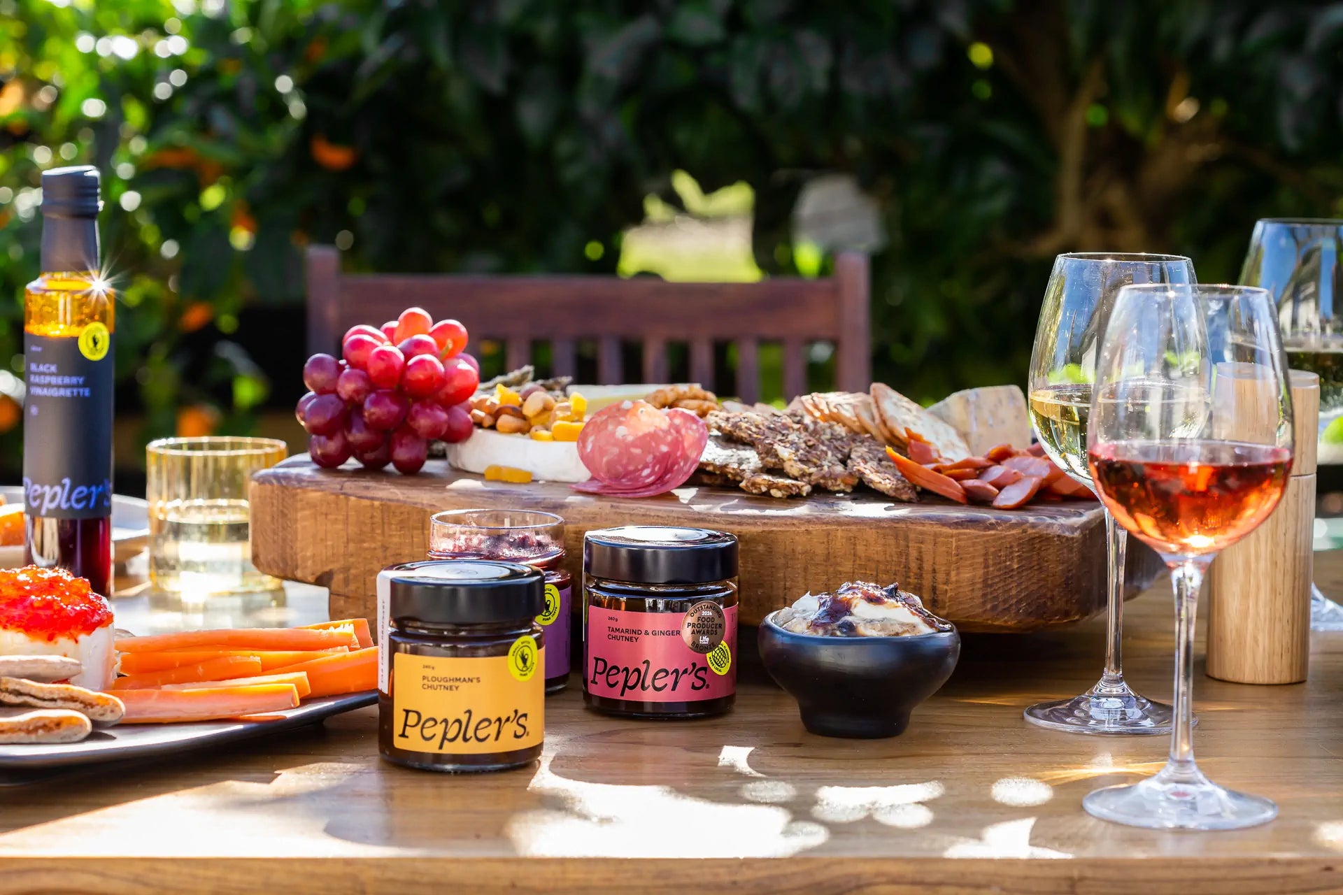 A beautifully arranged outdoor table setting featuring a wooden platter with a variety of artisanal cheeses, sliced meats, and nuts, complemented by a colourful assortment of grapes and dried fruits. In the foreground, jars of Pepler's chutneys are displayed, including 'Ploughman's Chutney' and 'Tamarind & Ginger Chutney.' Nearby, a small bowl holds a creamy dip topped with a sauce. Glasses of white and rosé wine sit beside an elegant oil bottle labeled 'Black Raspberry Vinaigrette.' The scene is set agains