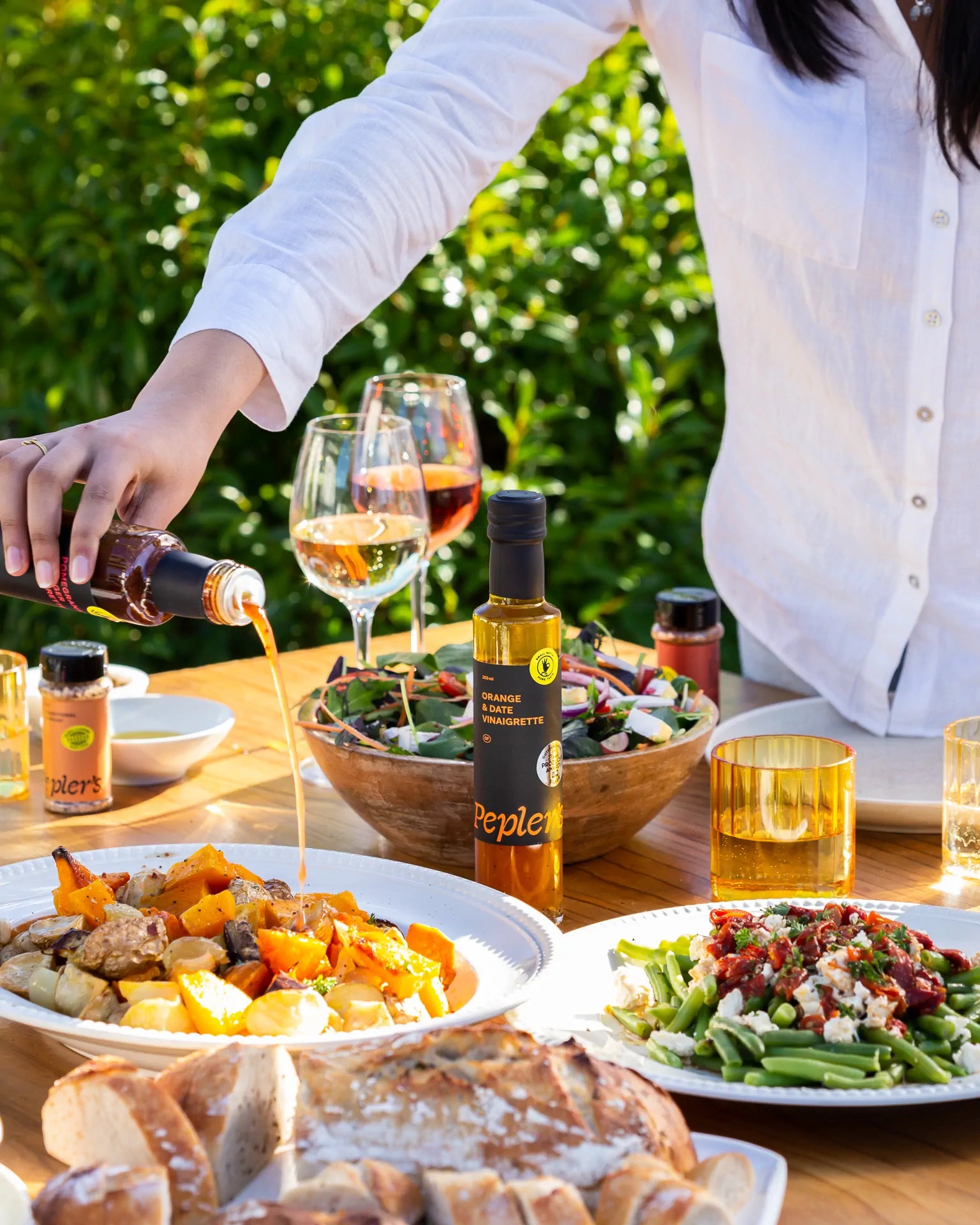 A hand pours a dark dressing from a bottle labeled "Pepler's" onto a colourful plate of roasted vegetables, a bottle of vibrant Pepler's orange and date vinaigrette is nearby, at an outdoor dining table. Nearby, a fresh salad in a wooden bowl, three glasses of wine, and dishes featuring green beans topped with sun-dried tomatoes and cheese are arranged alongside sliced artisanal bread. The scene is set in a lush, green garden with warm sunlight enhancing the inviting atmosphere of the meal.