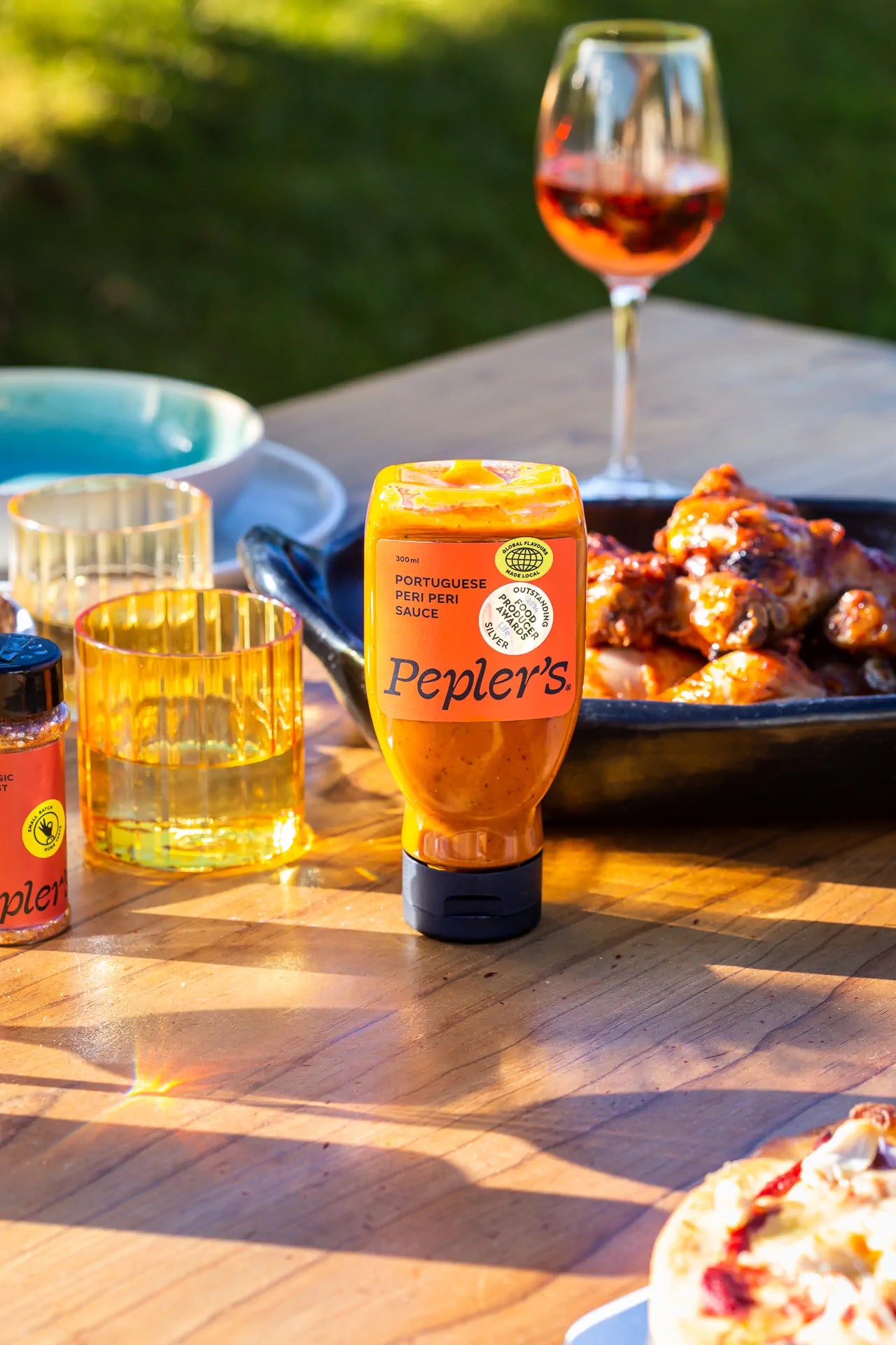 A bottle of Pepler's Portuguese Peri Peri sauce is prominently displayed on a wooden table, surrounded by a glass of rose wine, various decorative glasses, and a plate of glazed chicken wings. An additional spice container is visible, and a pizza slice can be seen at the edge of the table. The setting is bathed in warm sunlight, creating a vibrant outdoor dining atmosphere.