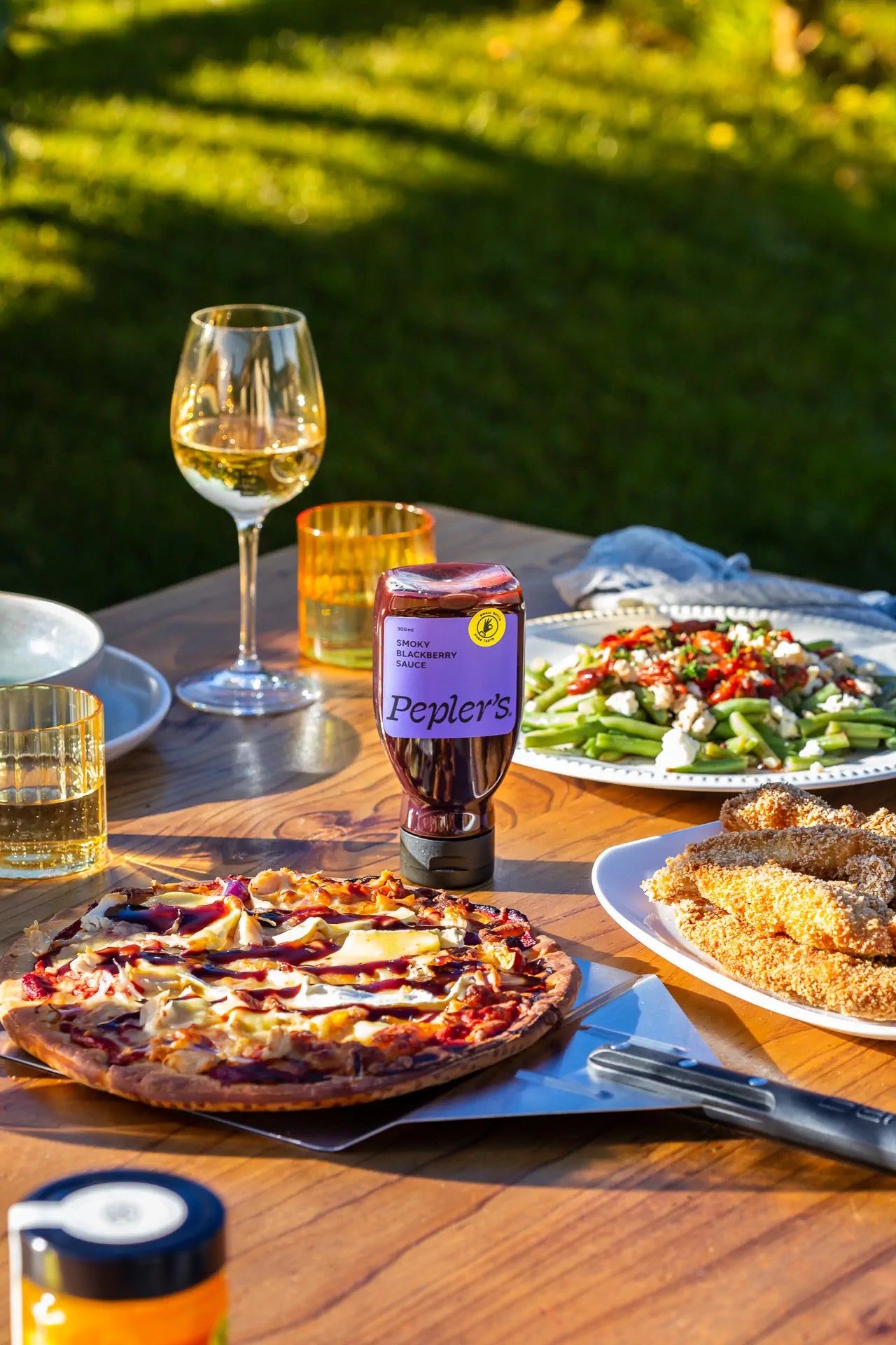 A wooden table set outdoors displaying a variety of dishes, including a round pizza topped with chicken and barbecue sauce, a plate of breaded chicken tenders, and a vibrant green bean salad garnished with red peppers and feta cheese. A bottle of Pepler's Smoky Blackberry Sauce is prominently featured in the center, alongside two glasses: one filled with white wine and another with a yellow drink. The background showcases lush green grass and soft sunlight, creating a warm and inviting atmosphere.