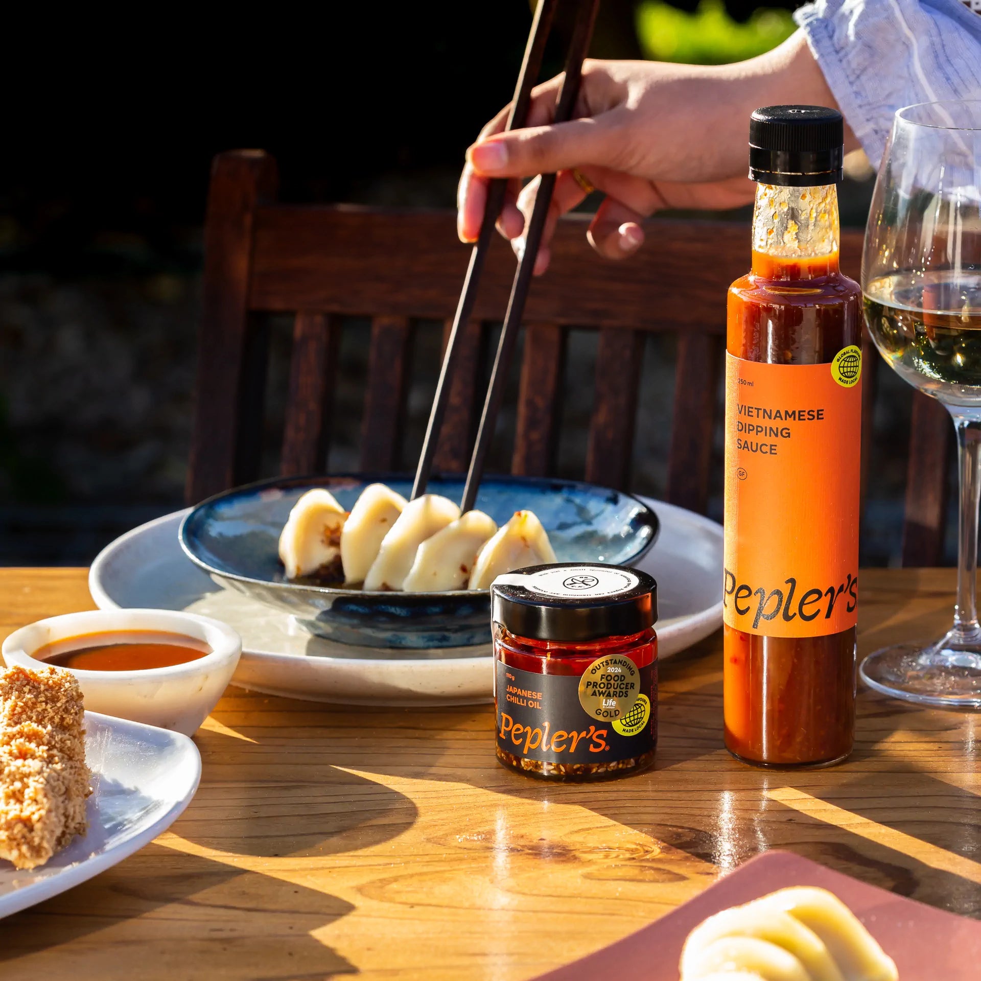 A hand using chopsticks to pick up dumplings from a blue plate, with a bottle of Pepler's Vietnamese Dipping Sauce and a jar of Pepler's Japanese Chilli Oil placed on a wooden table. In the background, a glass of white wine is visible, along with a piece of breaded food on a white plate. The scene is set outdoors, showcasing a well-lit and inviting dining experience.